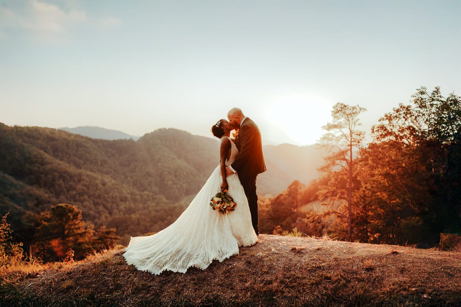 A bride and groom share a kiss at sunset, captured by a Smoky Mountain wedding photographer on a hill overlooking a lush, mountainous landscape. The brides bouquet complements her flowing dress, while the groom looks dapper in his suit among trees and distant hills bathed in warm sunlight.