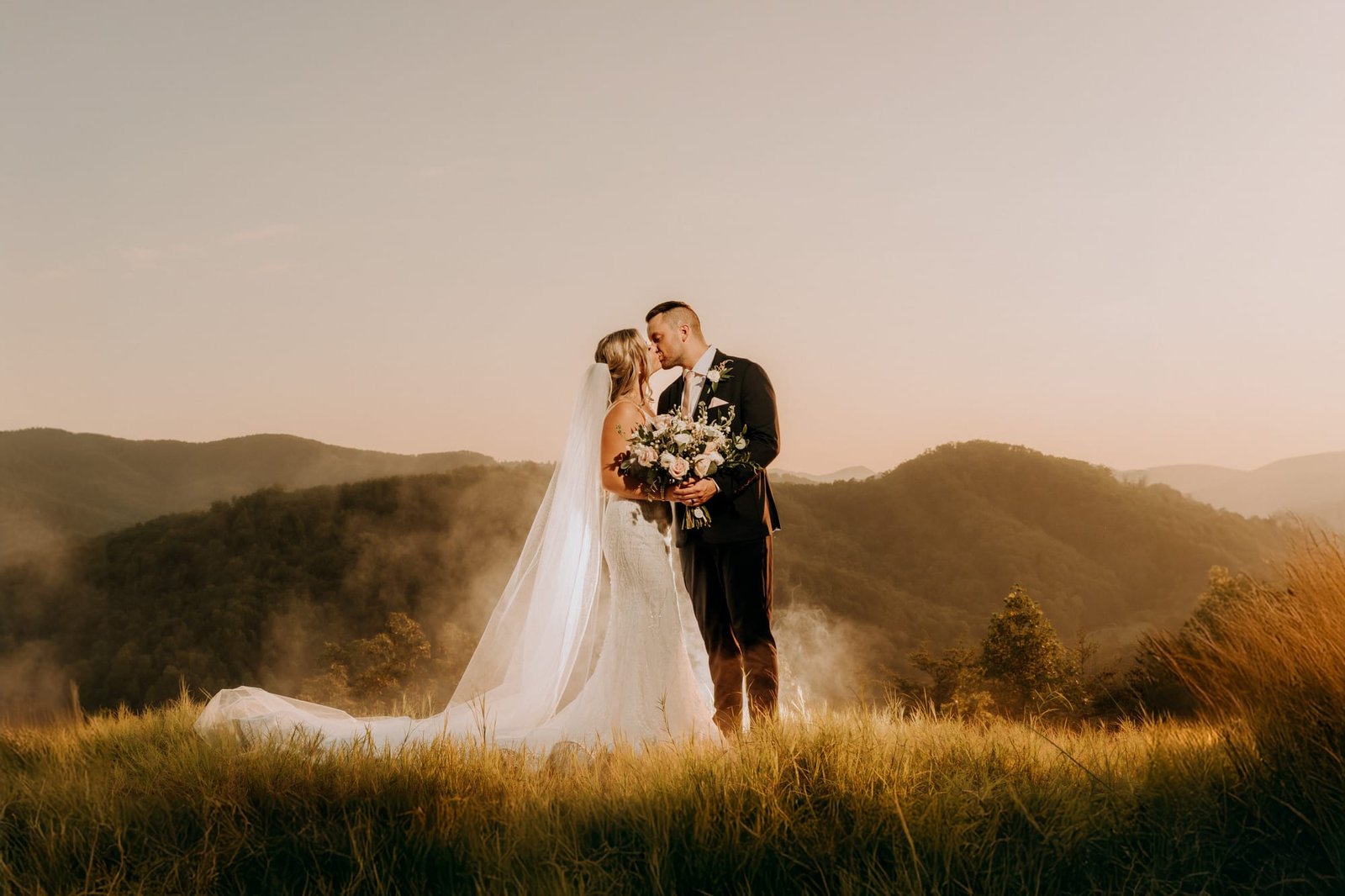 A bride and groom stand on a grassy hilltop with misty mountains in the background. The bride wears a long white gown and veil, and the groom is in a dark suit. They hold a bouquet of flowers and share an intimate moment at sunset.