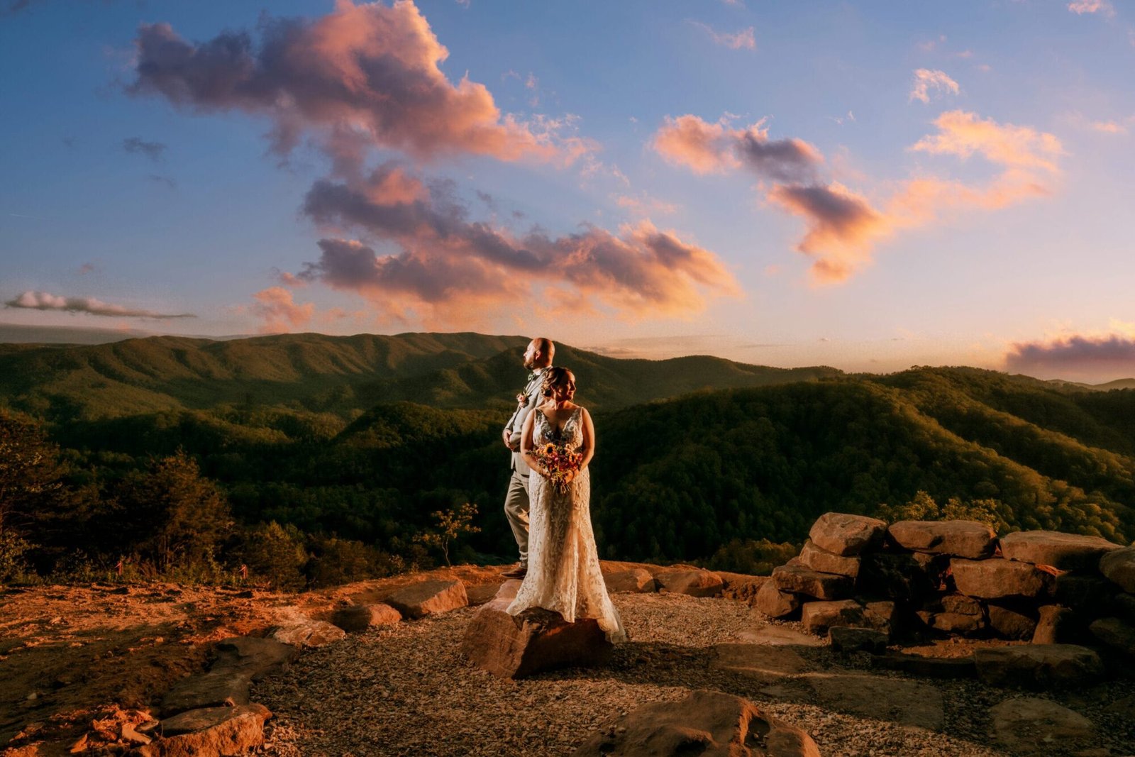 A bride and groom stand on a rocky summit, surrounded by a breathtaking mountain landscape at sunset. The sky is filled with pink and orange clouds, creating a romantic atmosphere.