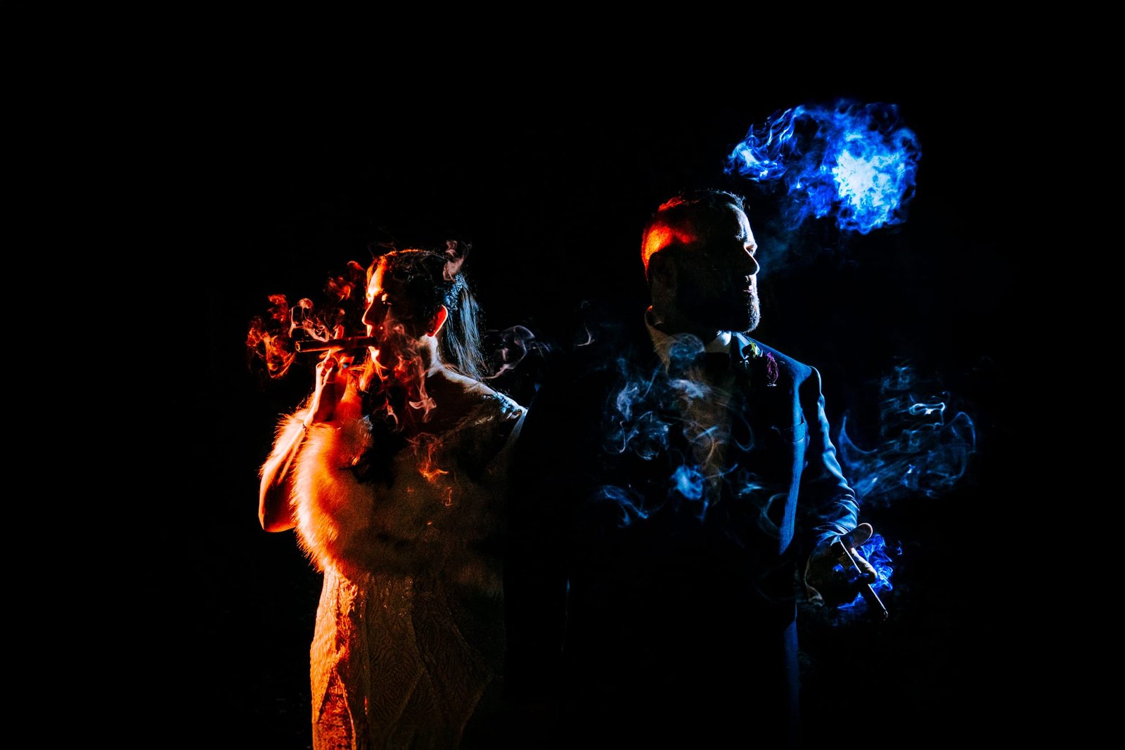 In a captivating scene captured by Derek Halkett Photography, a stylish couple stands enveloped in smoke. The woman, draped in a fur stole, exhales elegantly. Beside her, the man in a suit gazes aside as blue smoke swirls around them, contrasting vividly against the dark backdrop.