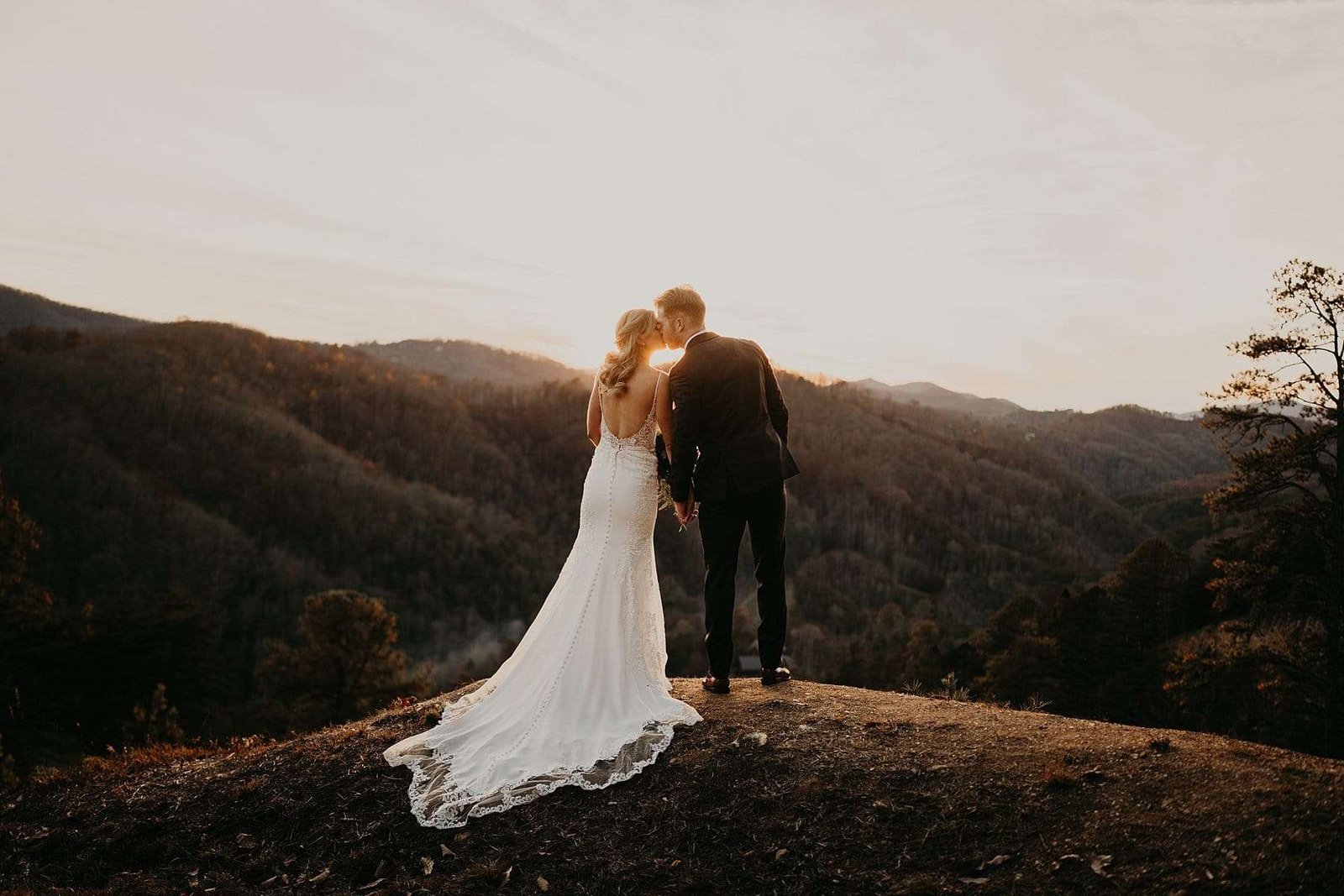 A couple in wedding attire stands on a hill at sunset, captured by Derek Halkett Photography. The woman in a white dress and the man in a dark suit face each other, overlooking a breathtaking mountainous landscape with trees and a partly cloudy sky.