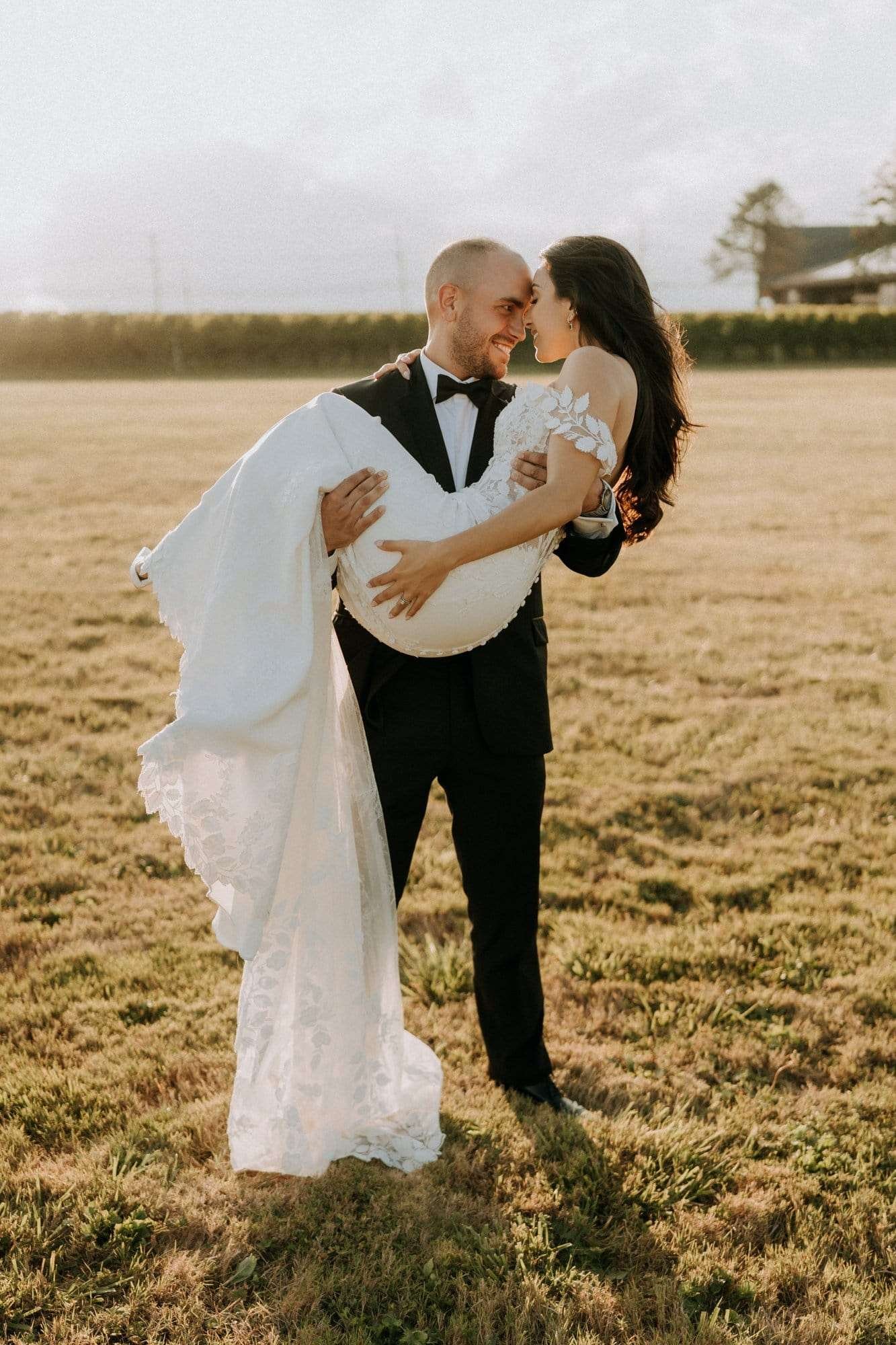 A groom in a black tuxedo holds a bride in a white gown in his arms. Captured by Derek Halkett Photography, they stand in a grassy field with the sun setting behind them. Both are smiling and leaning their heads close together, immortalizing this moment through an exceptional booking process.