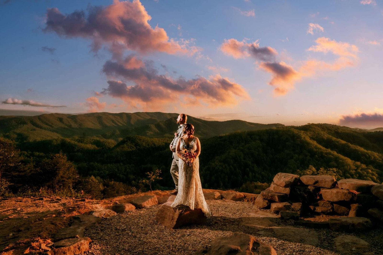 Derek Halkett Photography A bride and groom stand embracing on a rocky overlook during sunset, with a vast landscape of rolling hills in the Smoky Mountains and clouds in the background. The sky is a blend of oranges, pinks, and blues, creating a warm and romantic atmosphere. Smoky Mountain Wedding Photographer