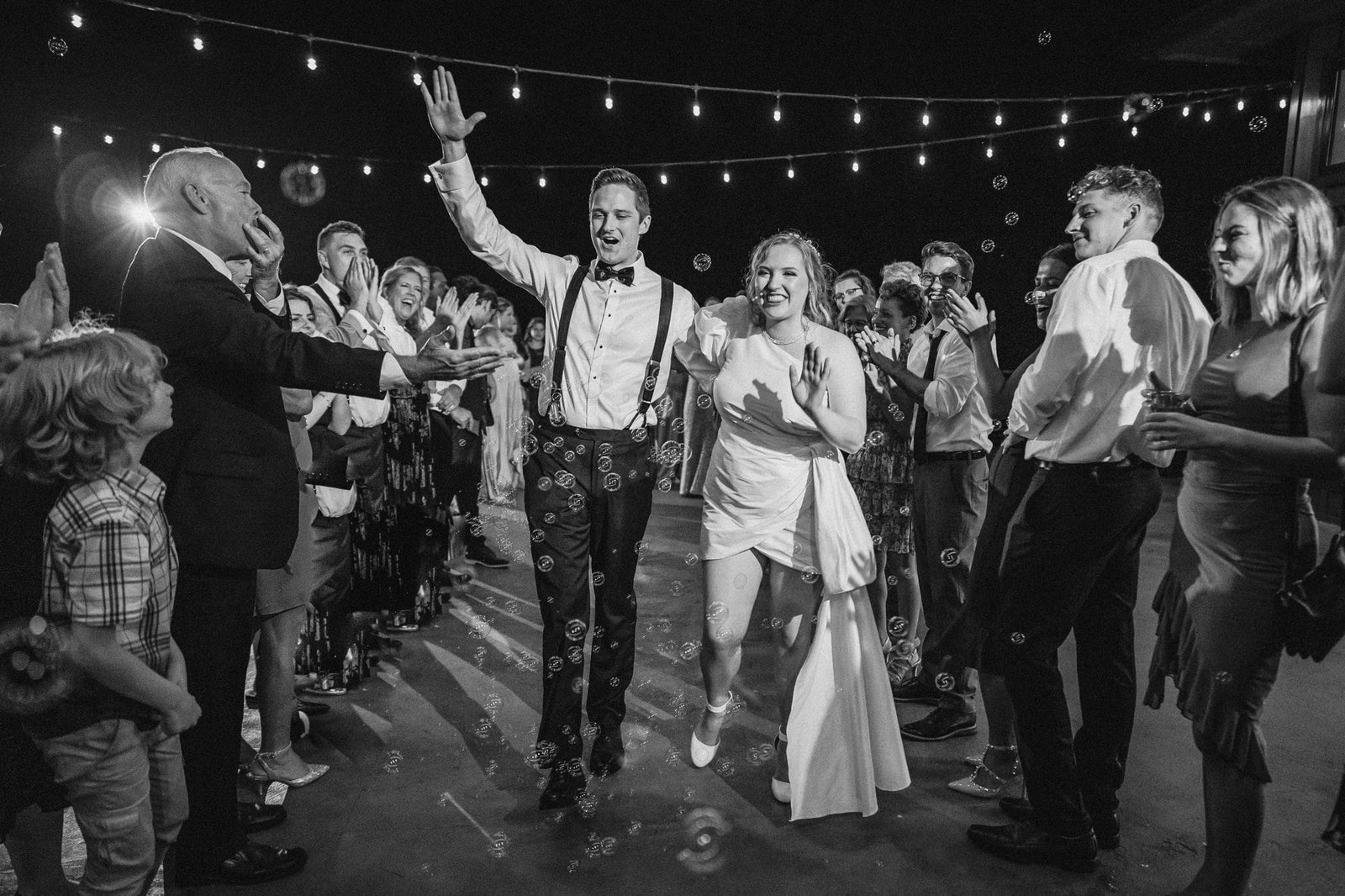 A black and white photo captures a joyful bride and groom illuminated by warm string lights, walking through a cheering crowd at night. The groom, stylish in suspenders, accompanies his bride in a white dress. Bubbles float around the couple as people clap and smile on both sides—a moment worthy of any wedding blog.