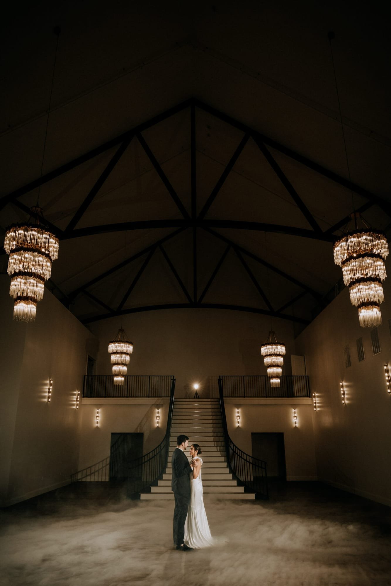 A couple embraces on a dimly-lit ballroom floor, captured in the distinctive style of Derek Halkett Photography. Illuminated by elegant chandeliers hanging from a high, vaulted ceiling, they create an enchanting scene reminiscent of a dreamlike social occasion.