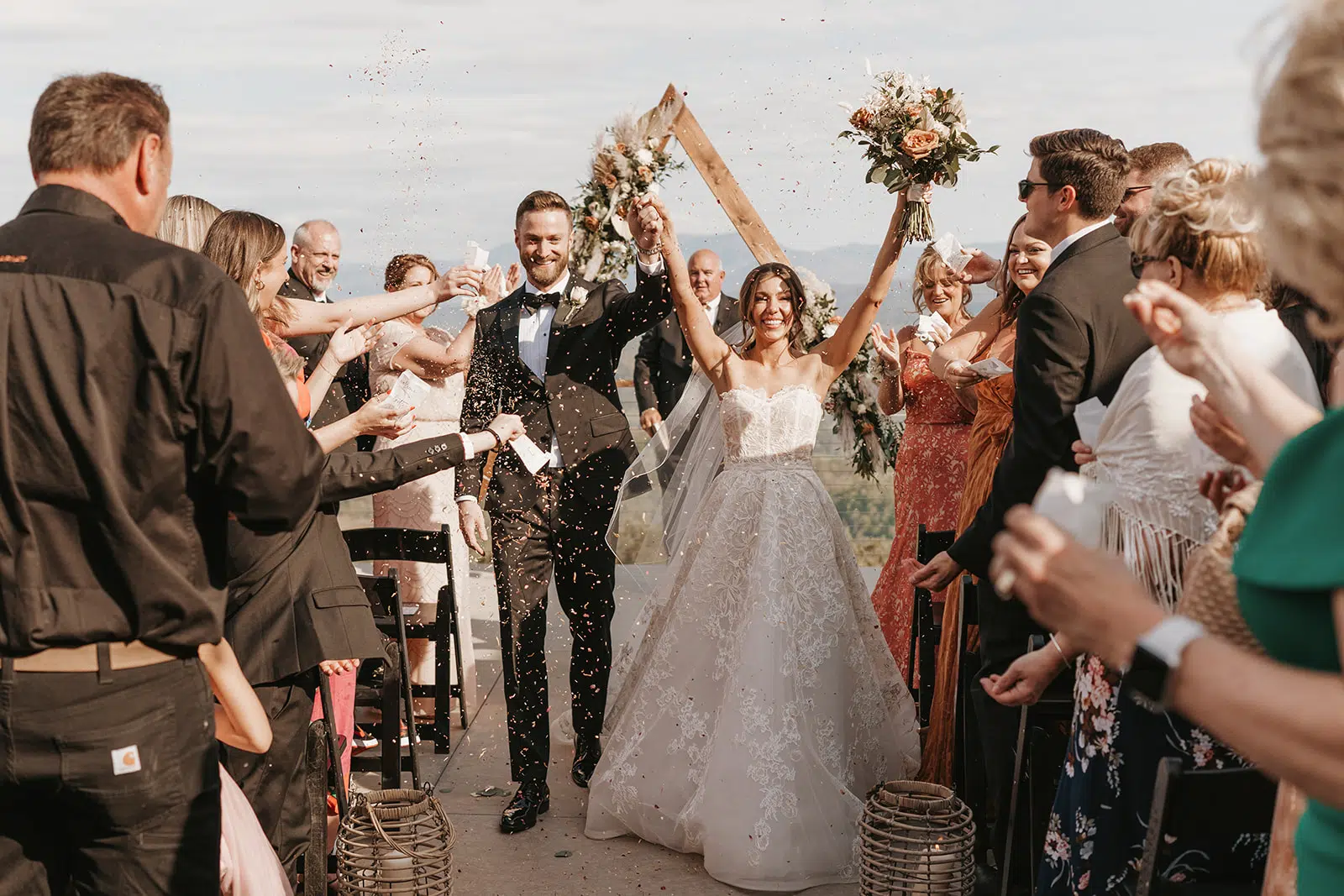 A joyful wedding scene unfolds with a bride and groom walking down the aisle, surrounded by cheering guests. The bride, in a white gown, holds flowers while the groom dons a suit. Confetti fills the air near a wooden arch adorned with blooms—a perfect capture for any Smoky Mountain Wedding Photographer.