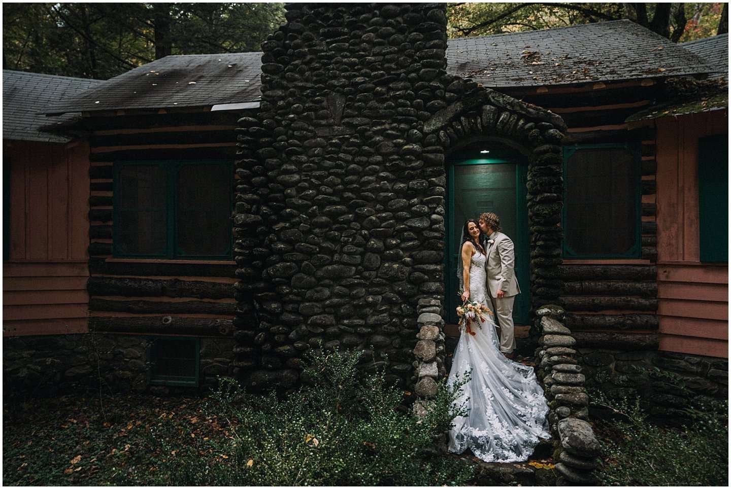 A bride and groom stand on a stone step in front of a rustic cabin, beautifully captured by a Smoky Mountain wedding photographer. The bride, in her long white gown, holds a bouquet, while the groom sports a light-colored suit. Trees surround the cabin, creating a peaceful, wooded setting.