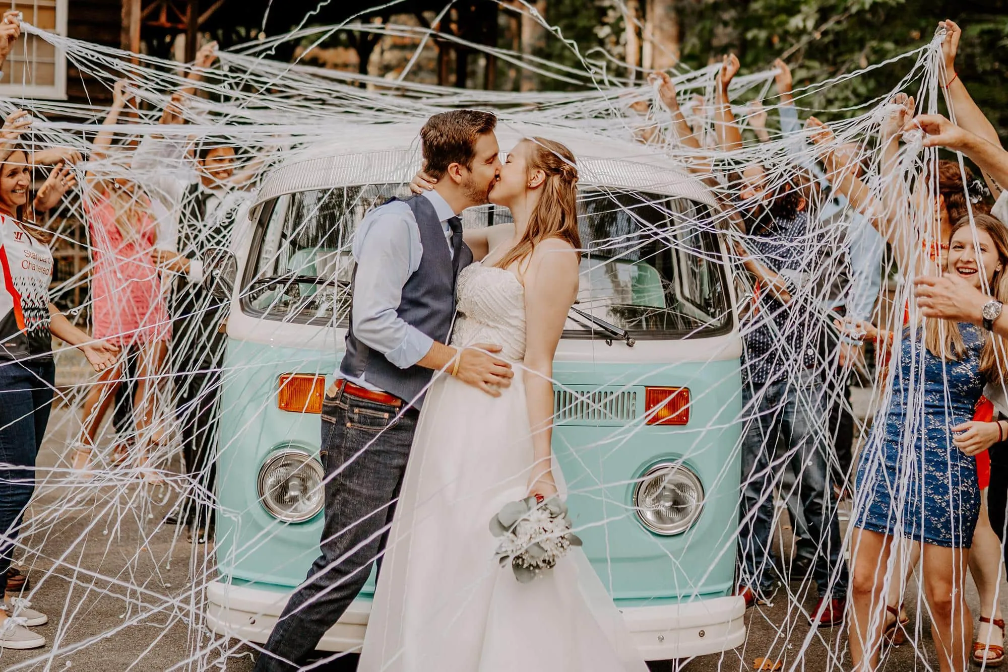 A bride and groom kiss in front of a vintage teal van, captured perfectly by their Smoky Mountain wedding photographer. Surrounded by cheering friends spraying strings of confetti, the scene is festive and joyful, sealing the celebratory moment of their big day.