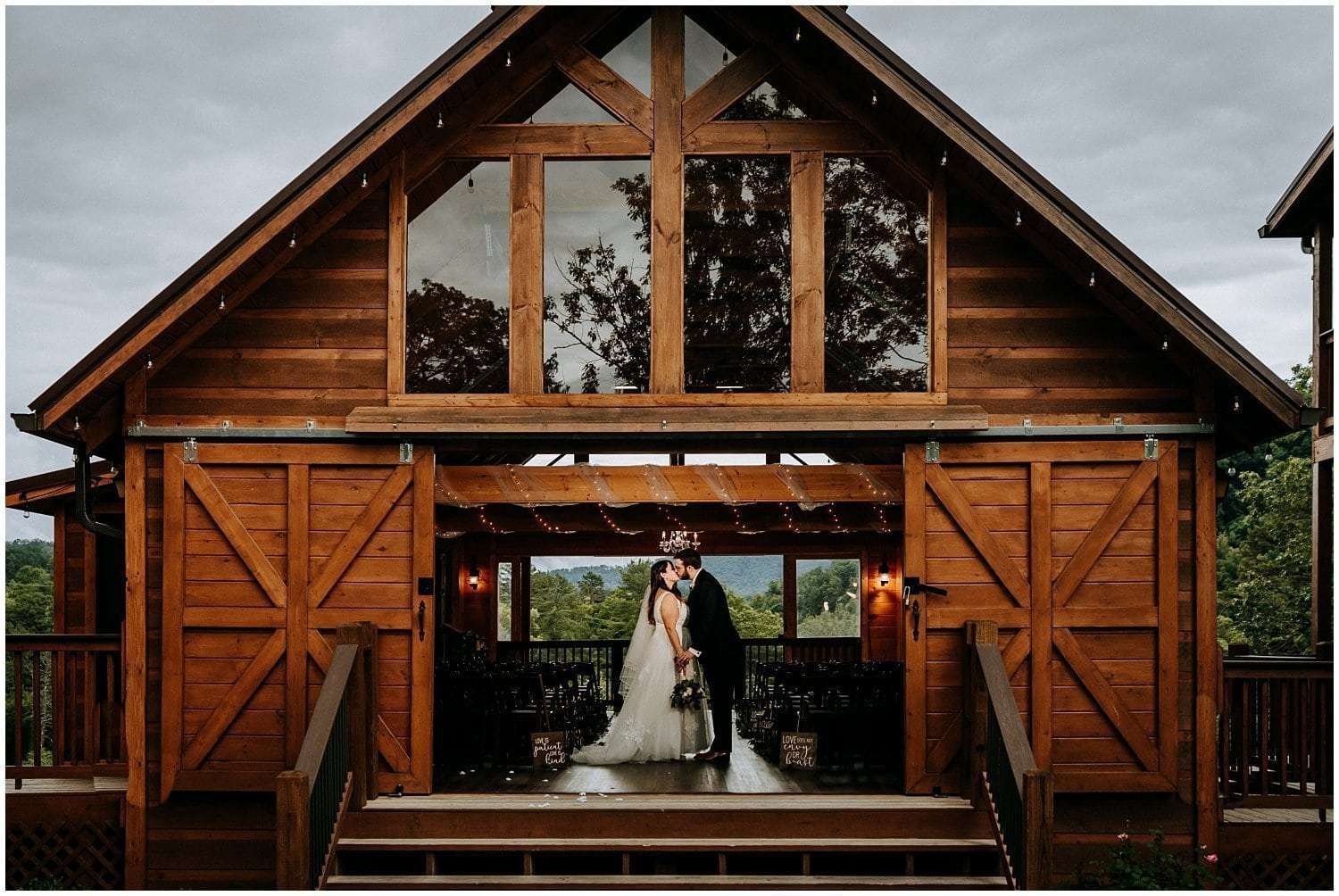 A couple in wedding attire stands on the wooden steps of a rustic barn. Captured beautifully by a Smoky Mountain Wedding Photographer, the large structure features glass details and is surrounded by trees, creating a romantic and picturesque setting.