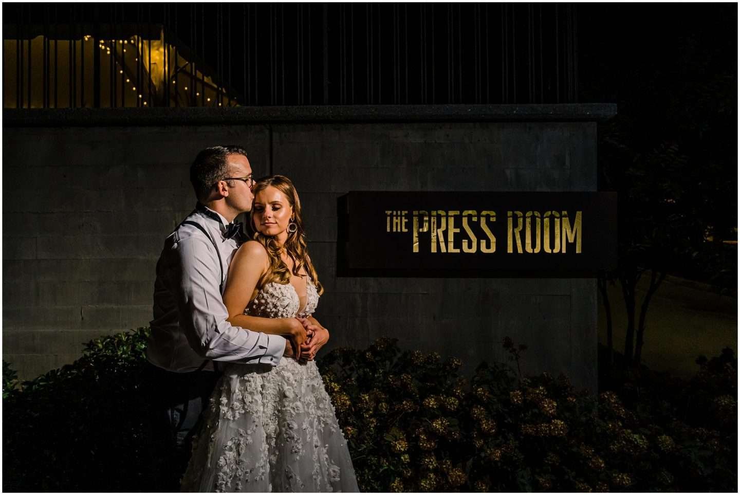 A couple embraces lovingly in front of The Press Room sign at night. The woman, in an elegant white gown, and the man, in a dress shirt, gently rest their heads together. Twinkling lights add a magical touch to this enchanting Press Room wedding scene.