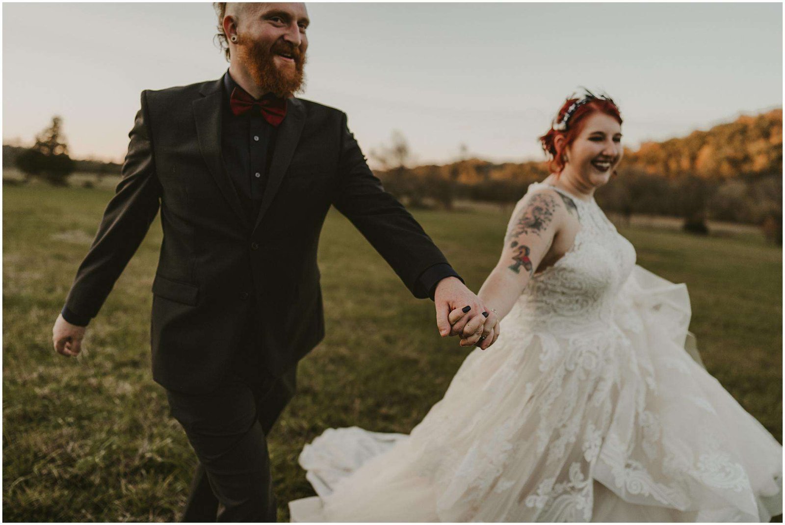 A couple, showcasing a wedding with distinction at The Barn at Chestnut Springs, are joyfully holding hands and walking through a grassy field. The person in a black suit and red bow tie is on the left, while the individual in a white lace gown adorned with visible tattoos is on the right.