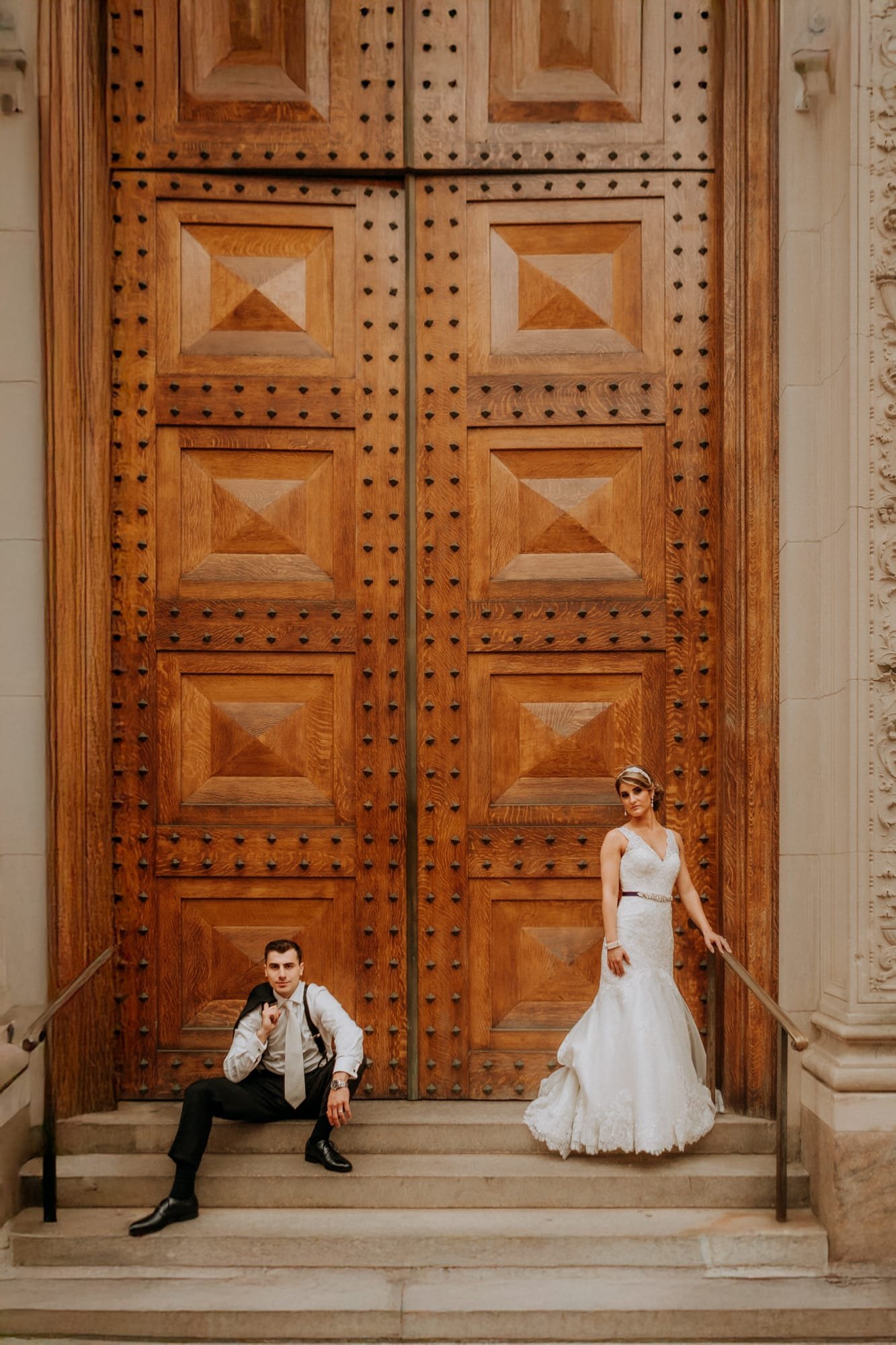 A couple in wedding attire poses in front of large, ornate wooden doors. Captured by a Smoky Mountain wedding photographer, the groom sits casually on the steps while the bride stands gracefully beside him, leaning against the door. The setting exudes elegance and architectural charm.