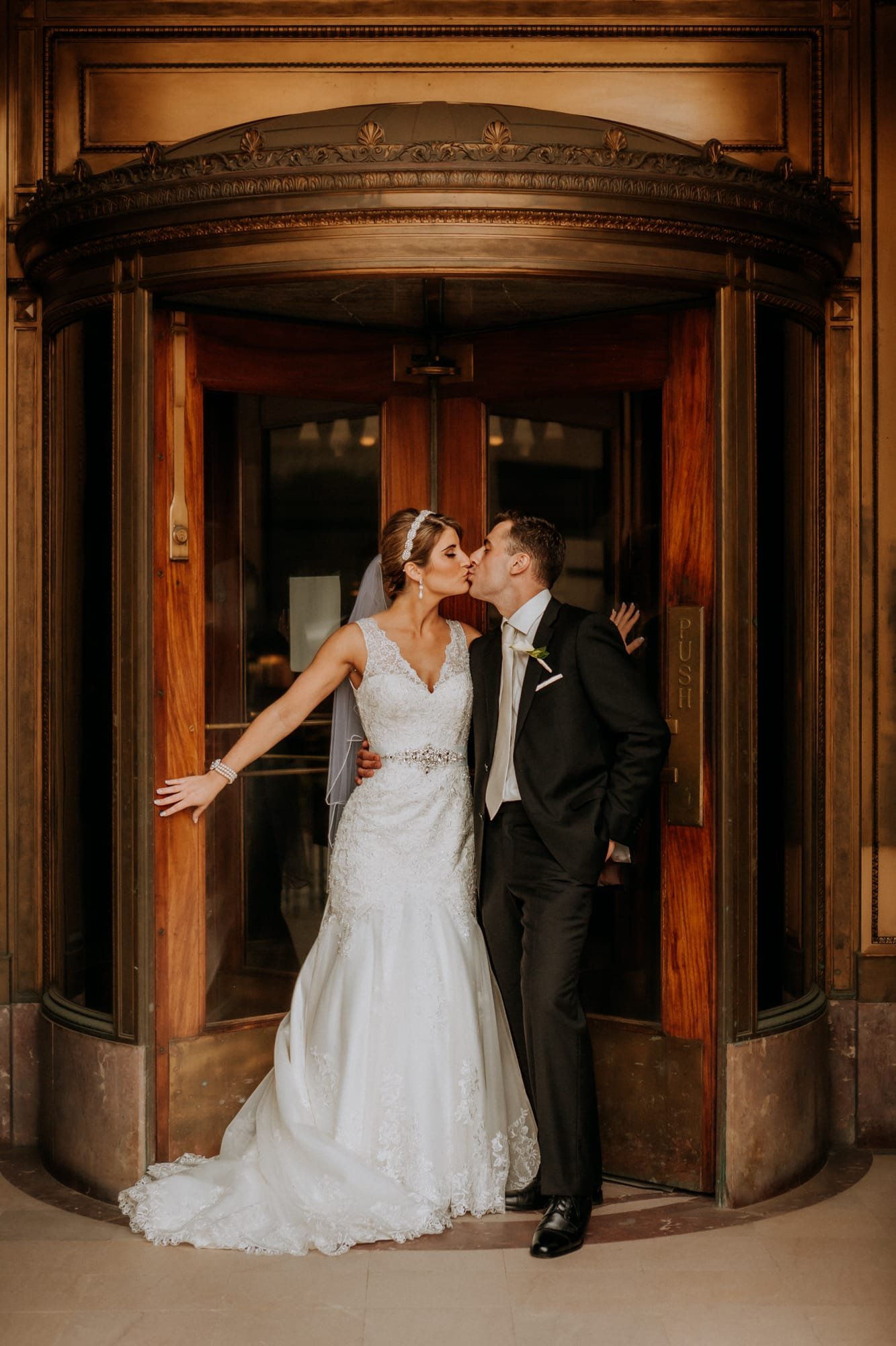 A bride and groom share a kiss in front of an ornate revolving door, captured flawlessly by a Smoky Mountain wedding photographer. The bride wears a white lace gown and veil, while the groom is in a black suit and tie, against the backdrop of an elegant building entrance.