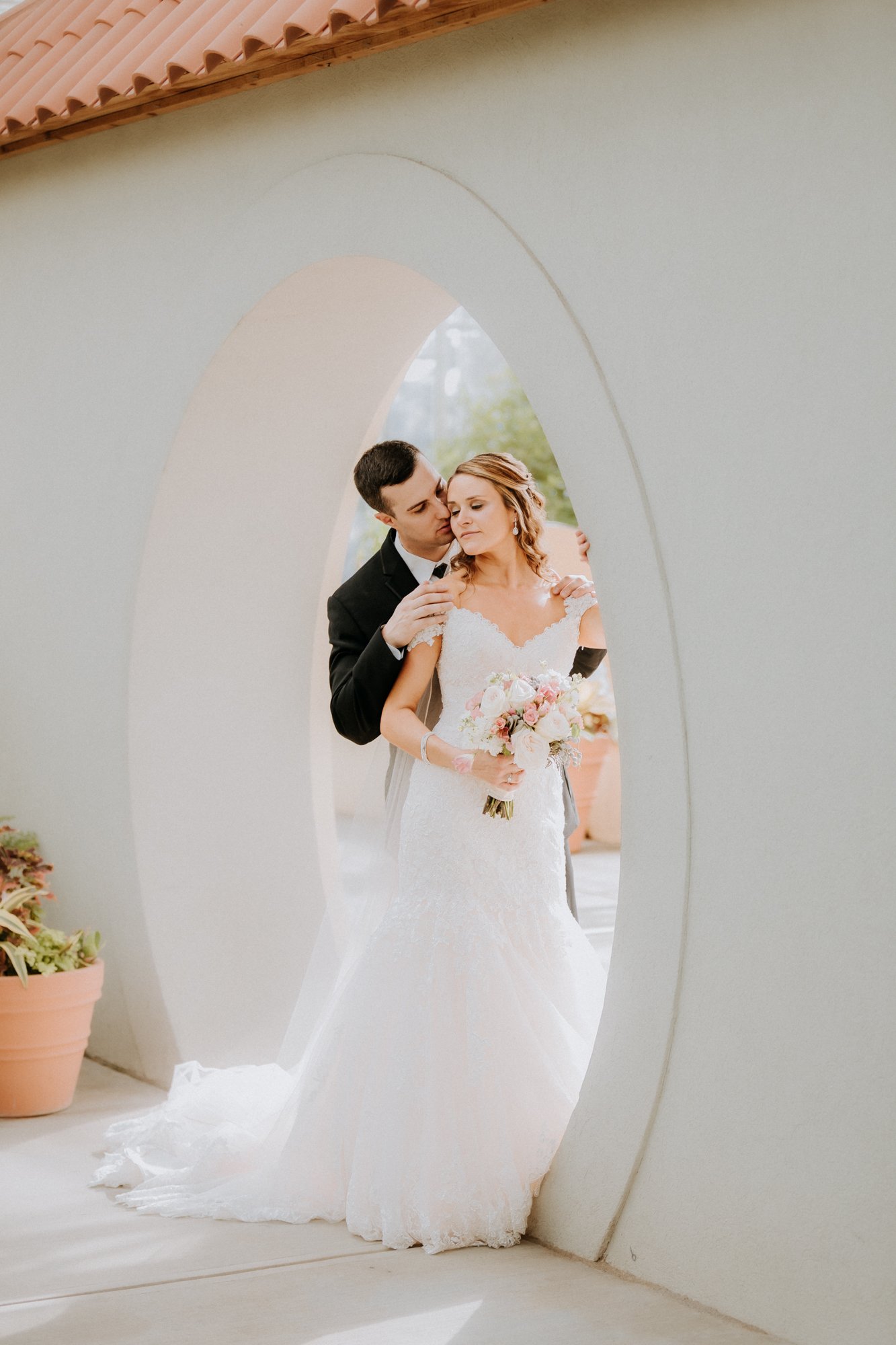 A couple poses for a photo captured by a Smoky Mountain wedding photographer on their special day. The bride, in a lace gown, holds a bouquet of flowers as the groom, in a suit, stands behind her. Theyre framed by a decorative archway with potted plants nearby.