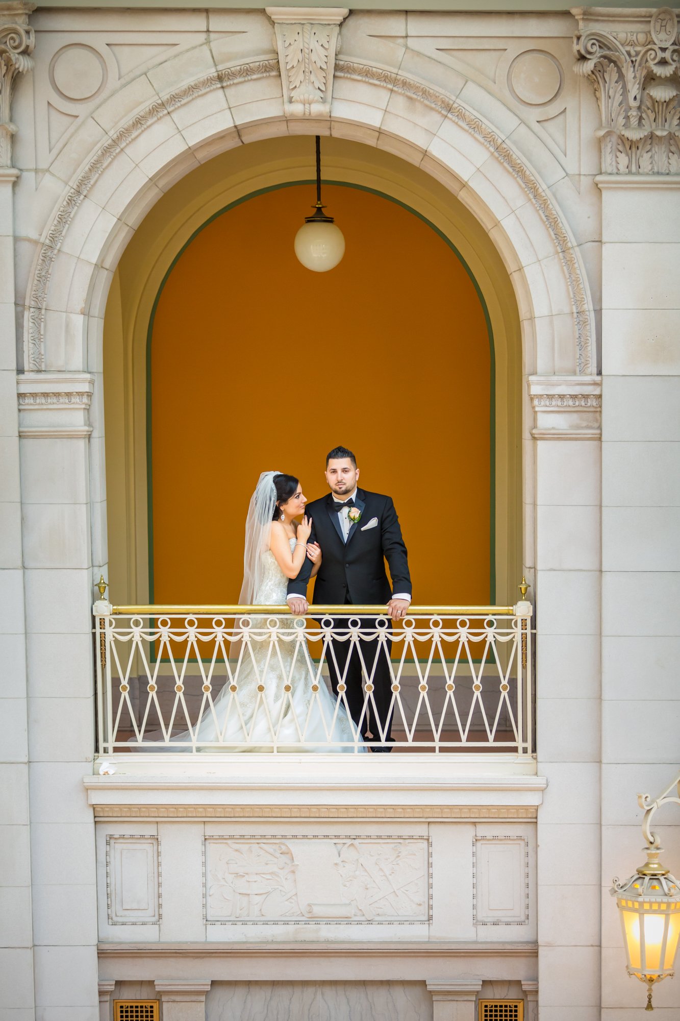 A bride in a white gown and veil stands next to a groom in a black suit and bow tie on a decorative balcony. Captured by a Smoky Mountain wedding photographer, they stand under an ornate archway with warm yellow-orange walls and a small hanging light above.