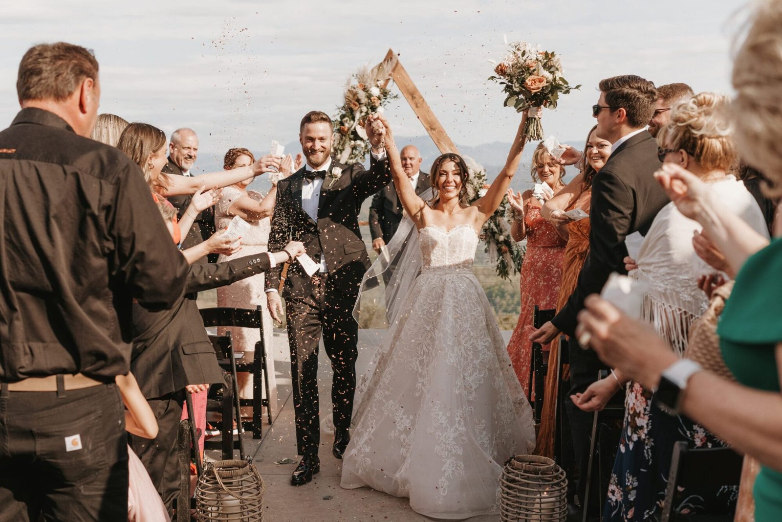 Amid the lush greenery of The Trillium Venue, a bride and groom walk down the aisle, smiling and holding hands. The bride raises her bouquet in celebration as guests cheer and toss confetti. An arch adorned with flowers completes this joyous scene for their Wedding Portfolio Page.