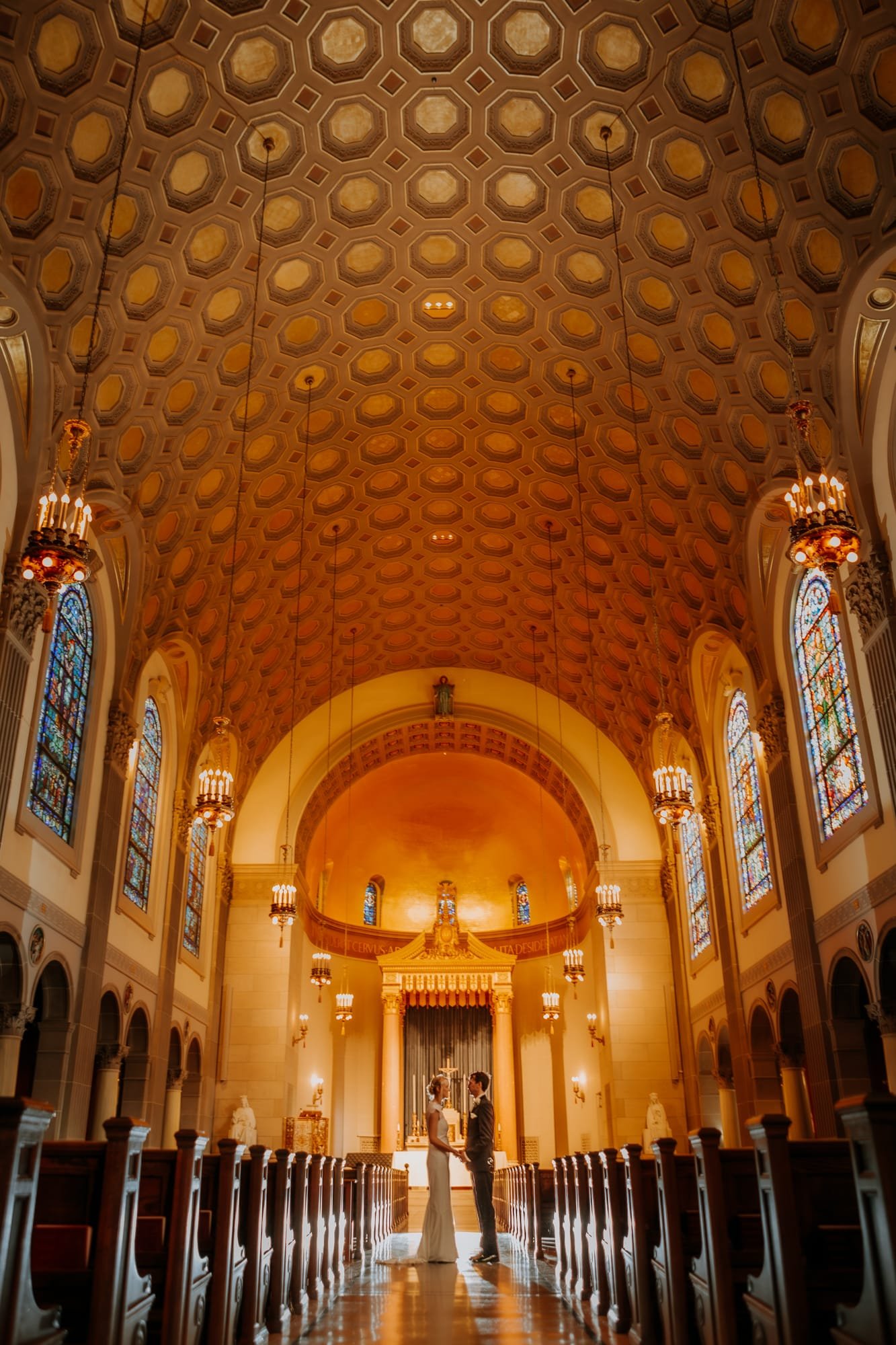 A couple stands in the aisle of a grand church under a high, ornate ceiling with geometric patterns. Stained glass windows line the walls, casting colorful light. Captured by a Smoky Mountain wedding photographer, they embrace in their wedding attire before the altar.