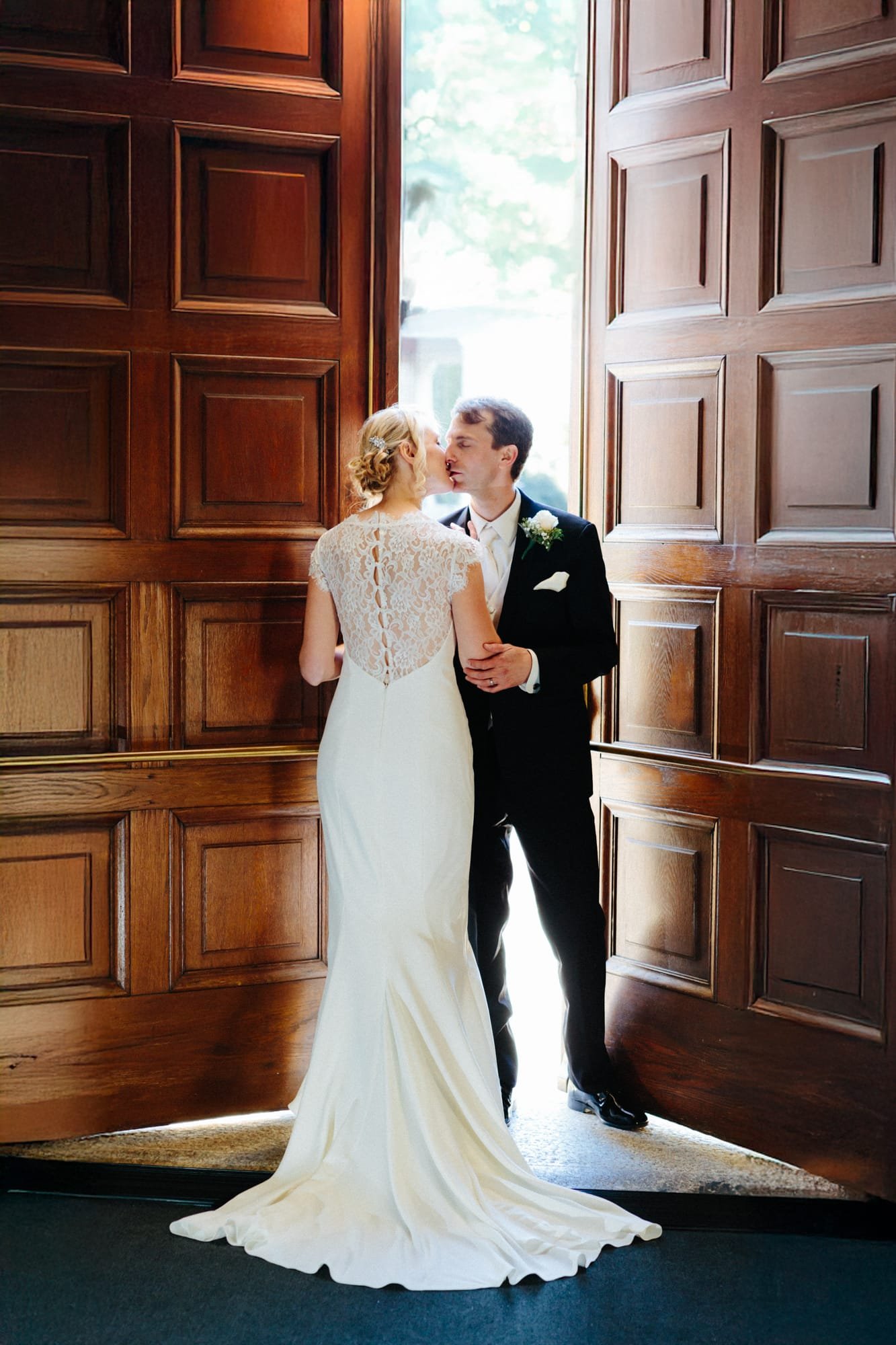 A bride and groom share a kiss at the entrance of a large wooden door, perfectly captured by their Smoky Mountain wedding photographer. The brides white lace gown and the grooms black tuxedo with a boutonniere are beautifully illuminated by natural light from outside.