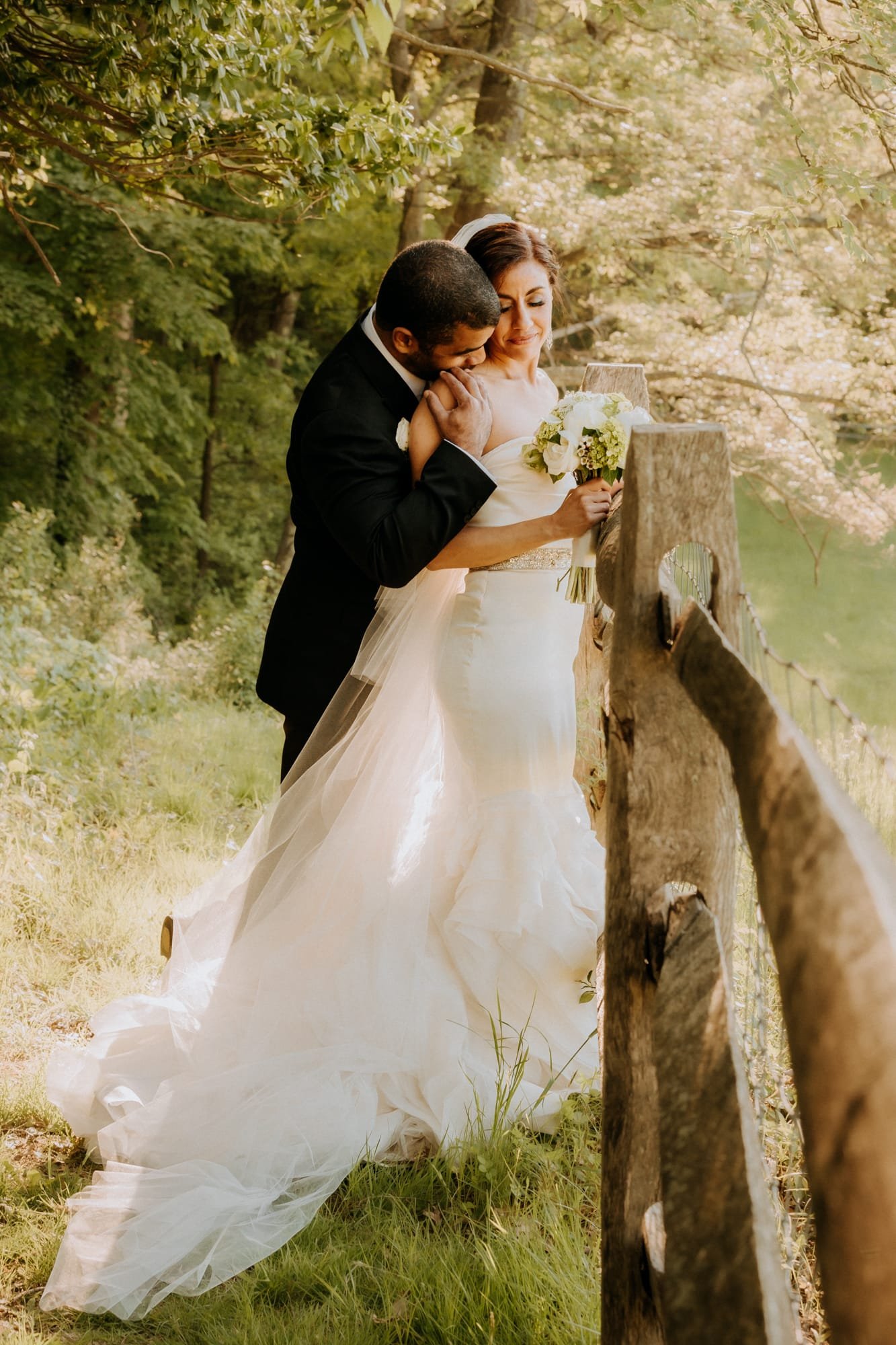 Bride and groom embrace beside a rustic wooden fence in a sunlit forest setting, captured by a Smoky Mountain wedding photographer. The bride holds a bouquet, and her long dress flows gracefully behind her as sunlight filters through the trees, creating a warm, romantic atmosphere.