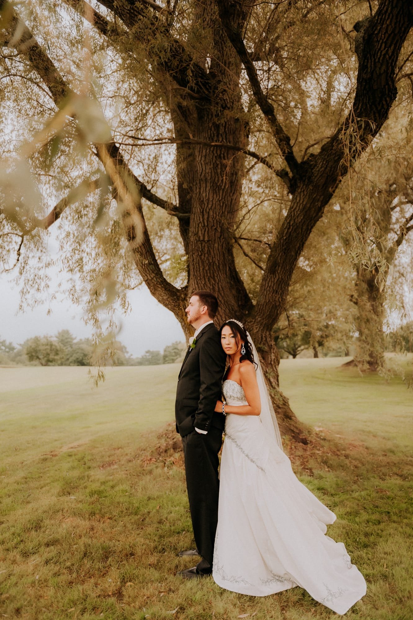 A bride and groom stand together under a large tree, captured beautifully by their Smoky Mountain wedding photographer. The bride, in a white dress and veil, leans her head on the grooms shoulder, who wears a black suit. The serene outdoor setting is enhanced by grass and additional trees.