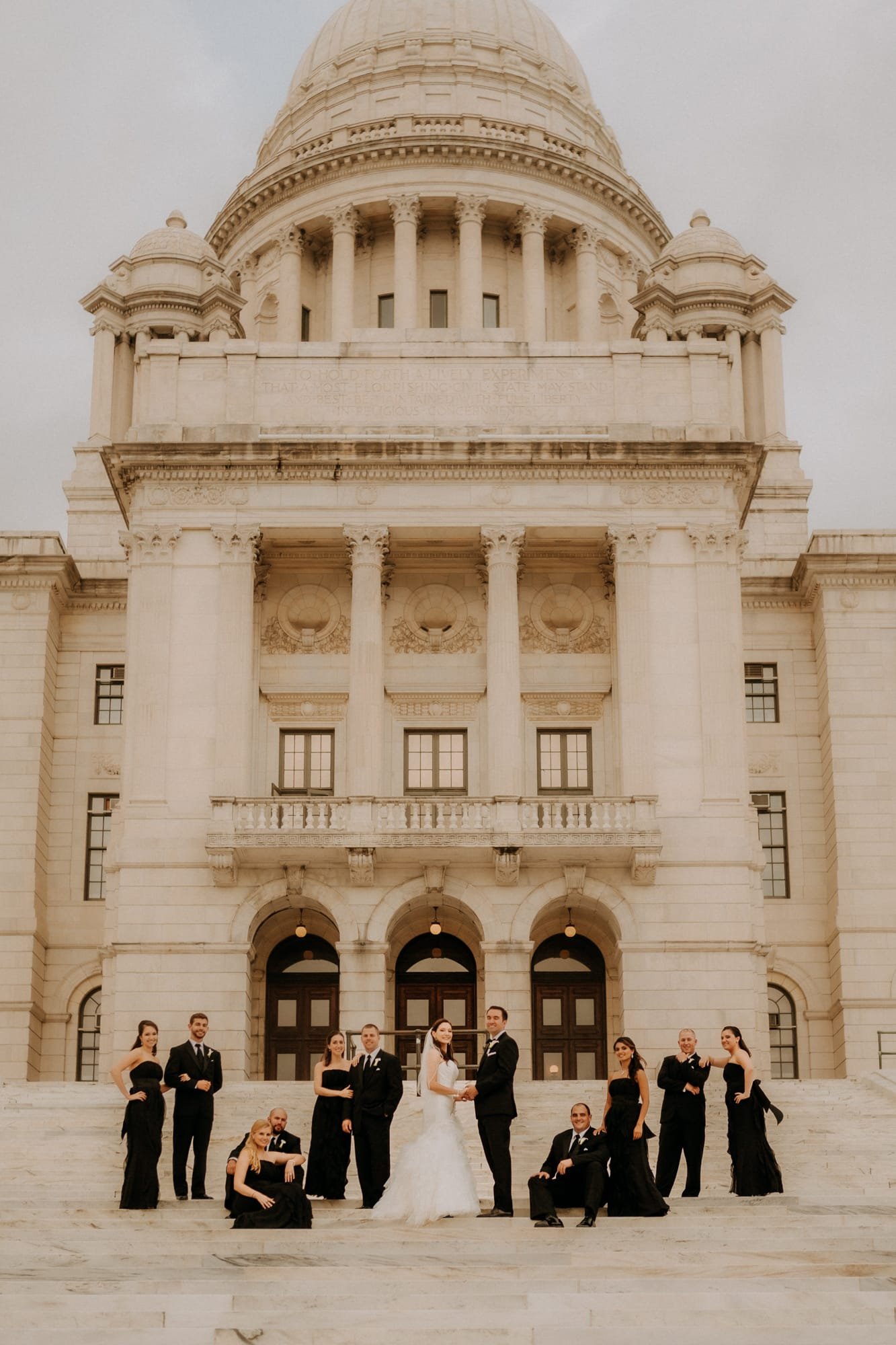 A bride and groom, captured by a Smoky Mountain Wedding Photographer, stand in front of a large stone building with a dome. Flanked by groomsmen and bridesmaids in black attire, the group poses on the steps, highlighting the buildings grand columns and intricate architectural details.