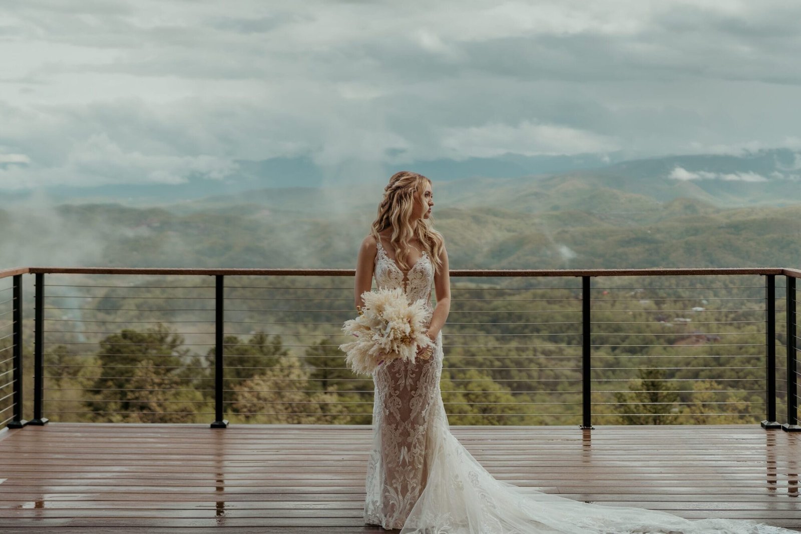 A bride in a lace wedding dress stands on a wooden deck at The Trillium Venue. She holds a bouquet of white flowers, gazing to the side against a backdrop of misty, rolling hills and a cloudy sky, capturing an unforgettable moment fit for any Wedding Portfolio Page.