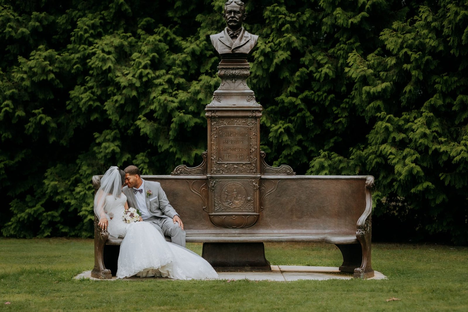 A bride and groom sit on a bronze bench under a statue bust, captured by a Smoky Mountain wedding photographer. The bride holds a bouquet as they share an intimate moment in the lush greenery, with tall trees creating a calm and romantic backdrop in the park.