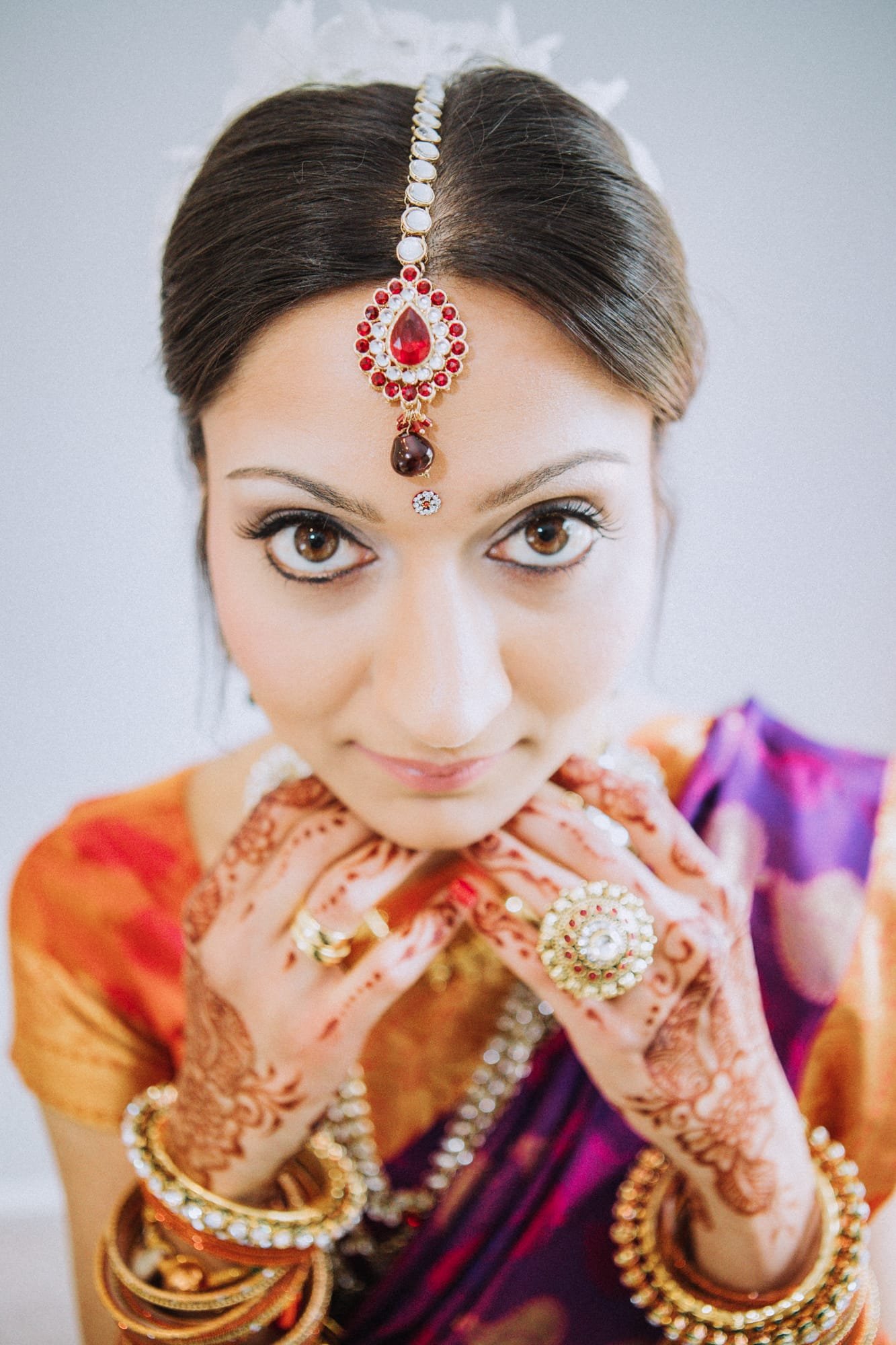 A woman in traditional attire gazes upwards, her colorful saree and intricate henna reflecting the artistry akin to a Smoky Mountain wedding photographer. Her jewelry, including a maang tikka, rings, and bangles, complements her serene expression and elegantly done makeup.