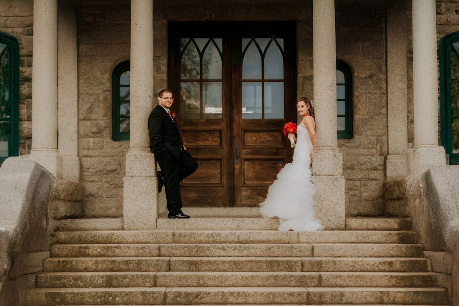 A bride in a white gown holds red flowers, standing on stone steps, while the groom in a black suit leans against a column. Captured by a Smoky Mountain wedding photographer, they stand outside a building with wooden doors and stone walls, surrounded by breathtaking natural beauty.